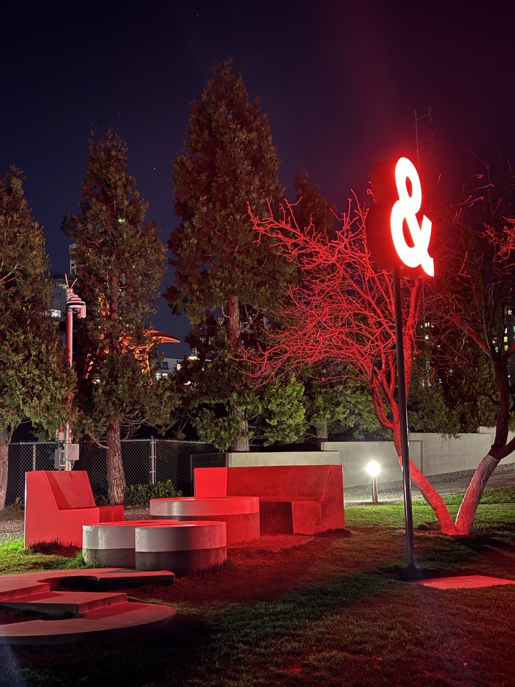 A glowing red ampersand sculpture lighting up trees and outdoor seating at night